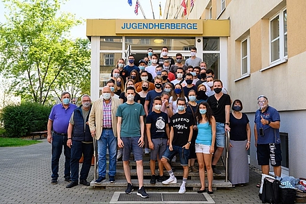 Gruppenfotos von Studierende auf einer Treppe vor einer Jugendherberge. Alle abgebildeten Studierenden tragen einen Mund-Nasen-Schutz.