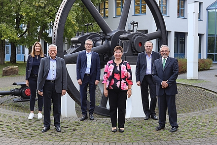 Sechs Menschen stehen vor einem schwarzen Schwungrad und lächeln in die Kamera. Sie stehen versetzt in zwei Reihen mit ca einem Meter Abstand zueinander. In der vorderen Reihe steht links ein grauhaariger älterer Mann mit schwarzer Hose, weißem Hemd und grauem Sakko. Neben ihm steht eine Frau im mittleren Alter. Sie trägt eine schwarze Hose und ein pink geblümtes Oberteil mit identisch assehendem Blazer. Neben ihr steht ebenfalls ein älterer Herr im Anzug. In der hinteren Reihe steht links eine blonde Frau in dunkler Kleidung. Neben ihr stehem zwei Herren in Anzüge. Im Hintergrund ist ein weißes Gebäude zu sehen.