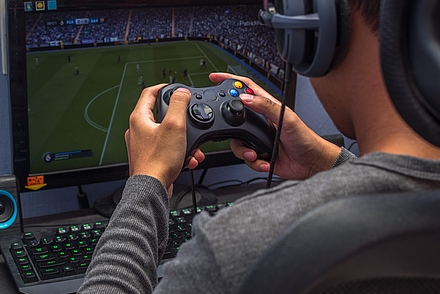 Person sitzt vor einem Bildschirm auf dem ein Fußballfeld zu sehen ist. Diese Person, die nur von hinten angeschnitten ist, trägt Kopfhörer und hält eine Konsole in der Hand.