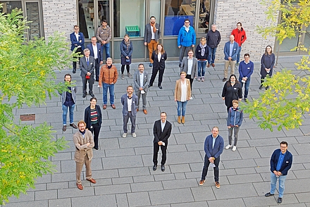 Fast 30 Menschen stehen mit abstand zueinander vor einem Gebäude und blicken lächelnd nach oben in dei Kamera. Links und rechts sind die grünen Blätter von zwei Bäumen zu sehen.