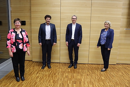 Vier Menschen stehen im Audimax mit einem Meter Abstand zueinander vor einer Holzwand. Links steht eine Frau im mittleren Alter, gekleidet in schwarzer Hose und pink geblümtem Oberteil mitsamt gleich aussehendem Blazer. Neben ihr steht ein Herr mit dunklem Lockenkopf in Anzug. Daneben steht ein grauhaariger Mann ebenfalls in Anzug. Ganz rechts steht eine blonde Frau, ebenfalls mittleren Alters, die eine dunkel Hose, ein blau geblümtes Obertel und einen blauen Blazer trägt. Alle vier lächeln sie in die Kamera.