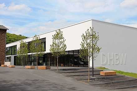 Das E-Gebäude am Campus Mosbach. Das Gebäude ist ein weißer, rechteckiger Neubau mit einem Flachdach und einem DHBW Schriftzug an der rechten Außenwand. Vor dem Gebäude sieht man den Hof.