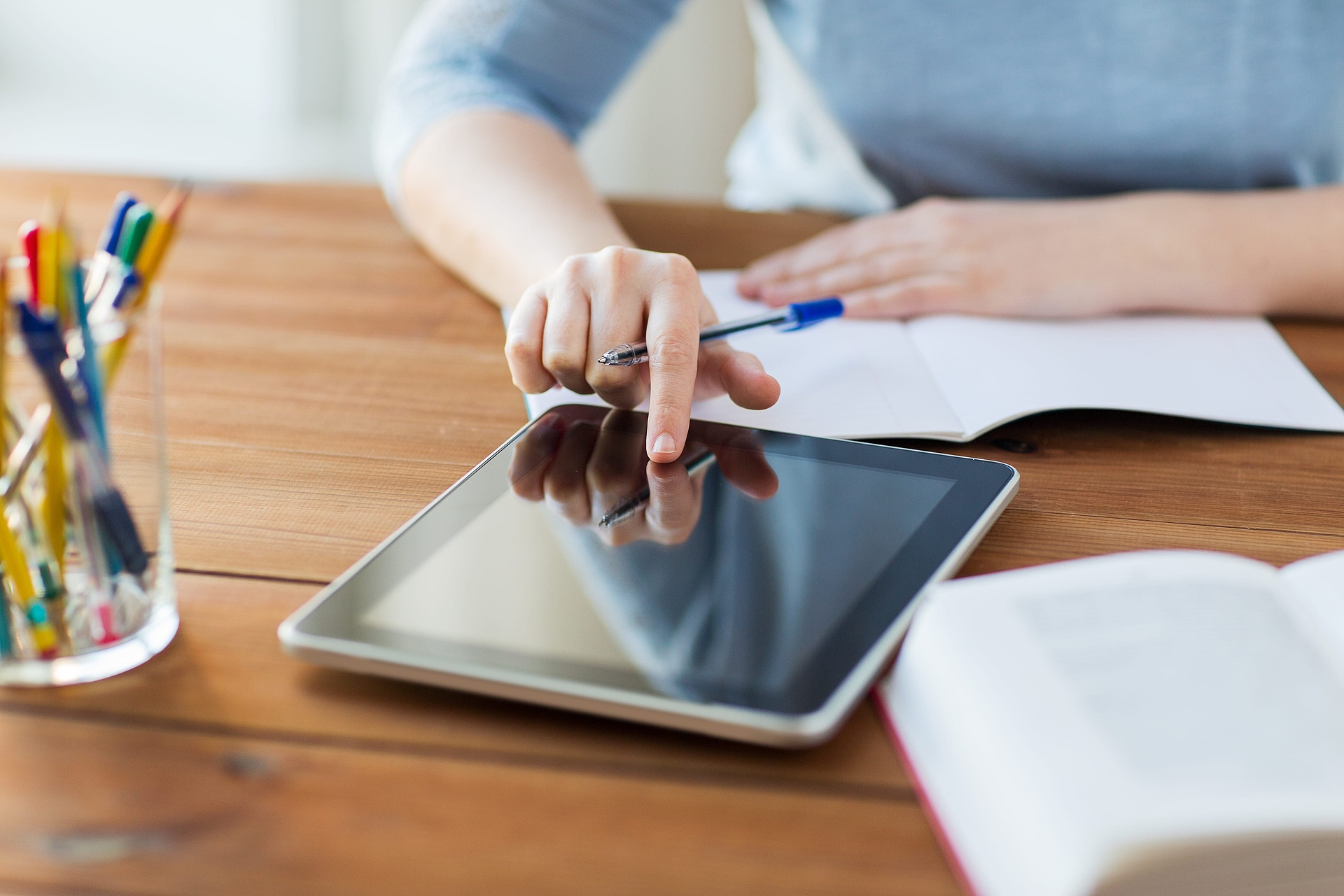 Ein Tablet liegt auf einem Holztisch. Man sieht einen Ausschnitt eines Oberkörpers mit hellblauen Oberteil, welcher an diesem Tisch sitzt und auf das Tablett tippt. Vor der Person auf dem Tisch liegt zudem ein weißes Papier. Neben dem Tablett steht ein Glas voller Stifte und ein aufgeschlagenes Buch.