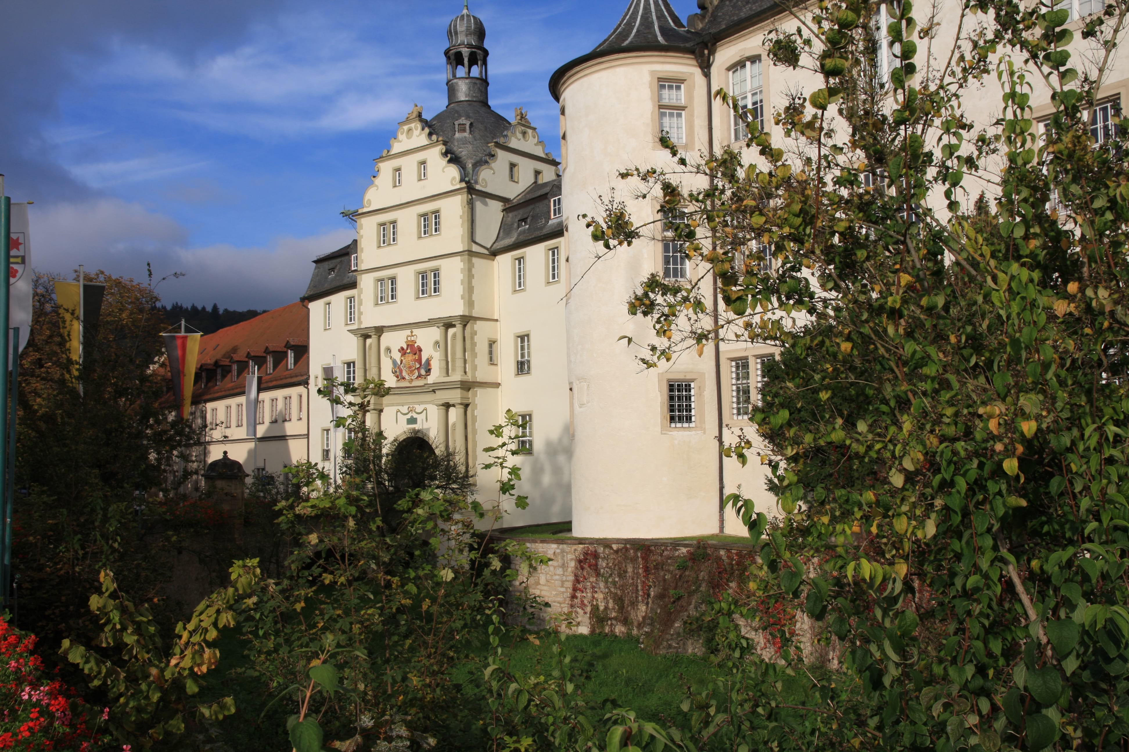 Auf dem Bild ist ein Ausschnitt des Deutschordenschlosses in Bad Mergentheim zu sehen. Das helle Schloss ist im Ausschnitt von dunkelgrünen Büschen umgeben. Der Himmel strahlt im satten blau, ein paar leichte Wolken sind zu sehen.