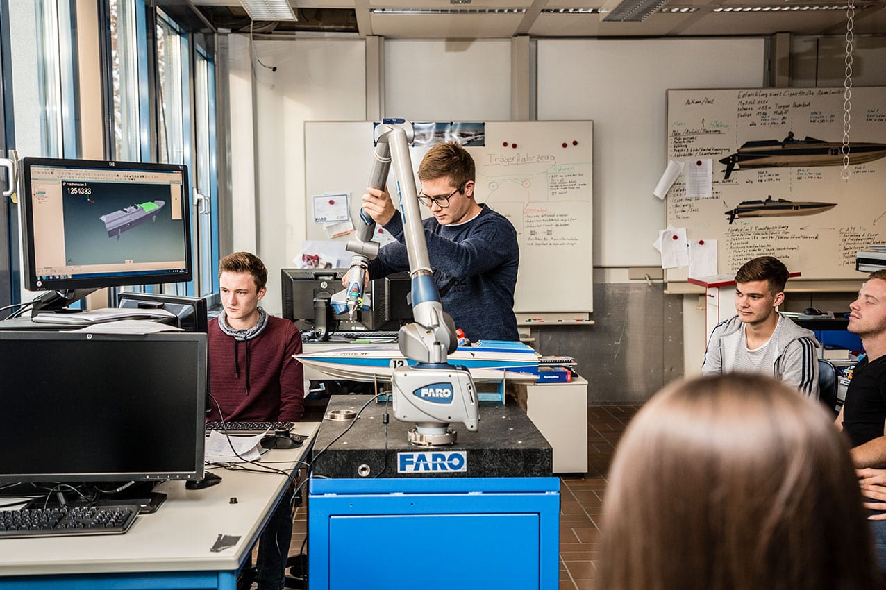 Ein junger Student, mit dunkelblonden kurzen Haaren, einer schwarzen Brille und dunkelblauen Pulli steht mittig vor einem Roboterarm und bewegt diesen. Rechts im Bild sitzen Studierende auf Stühlen und schauen dem anderen zu. Links im Bild sitzt ein weiterer Student mit weinrotem Sweatshirt vor mehreren Bildschirmen. Die Szene findet in einem Kursraum mit zwei Memoboards und einer Tafel statt.