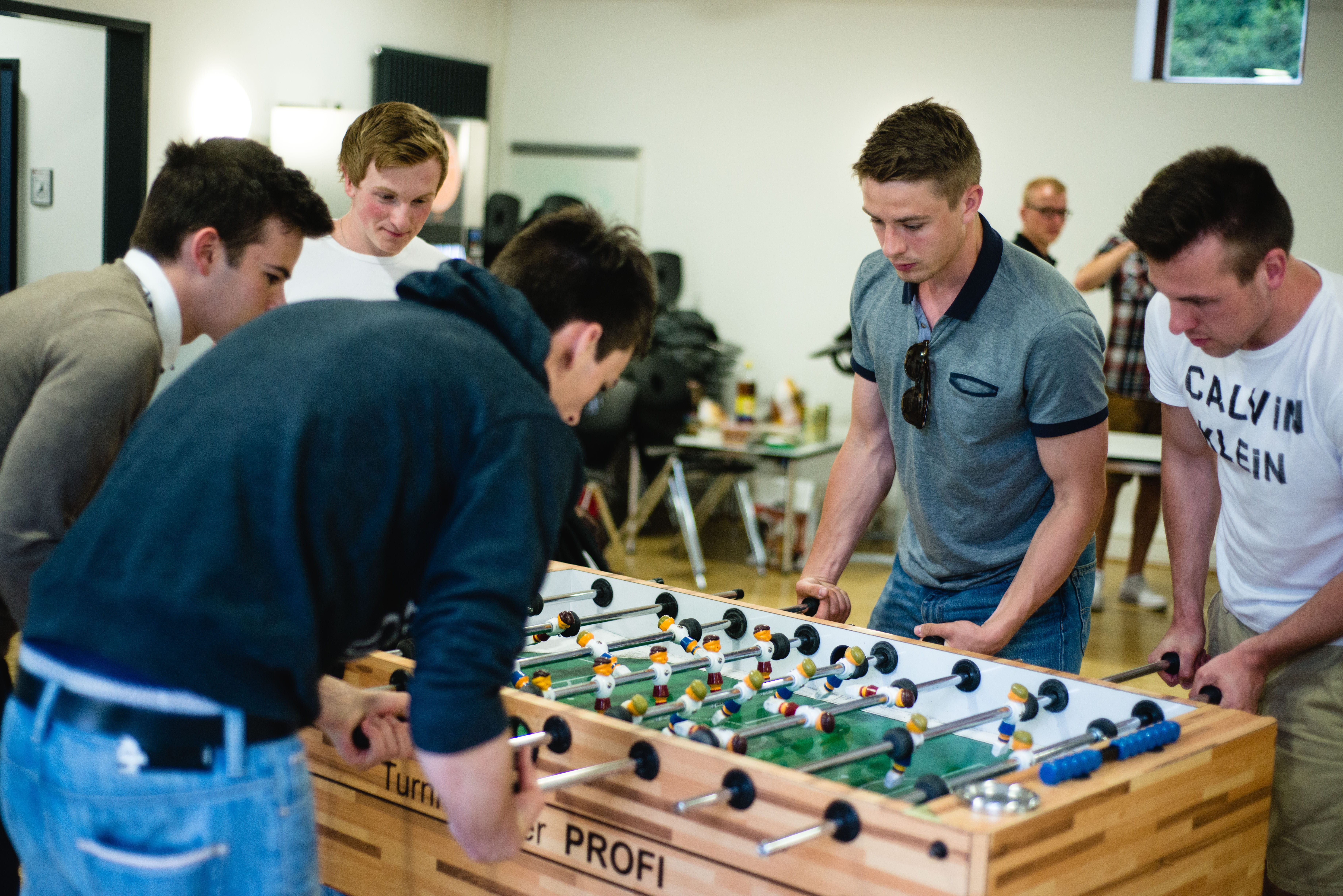 Um einen Tischkicker stehen fünf Studenten. Vier spielen in zweierteams gegeneinander, einer schaut zu. 