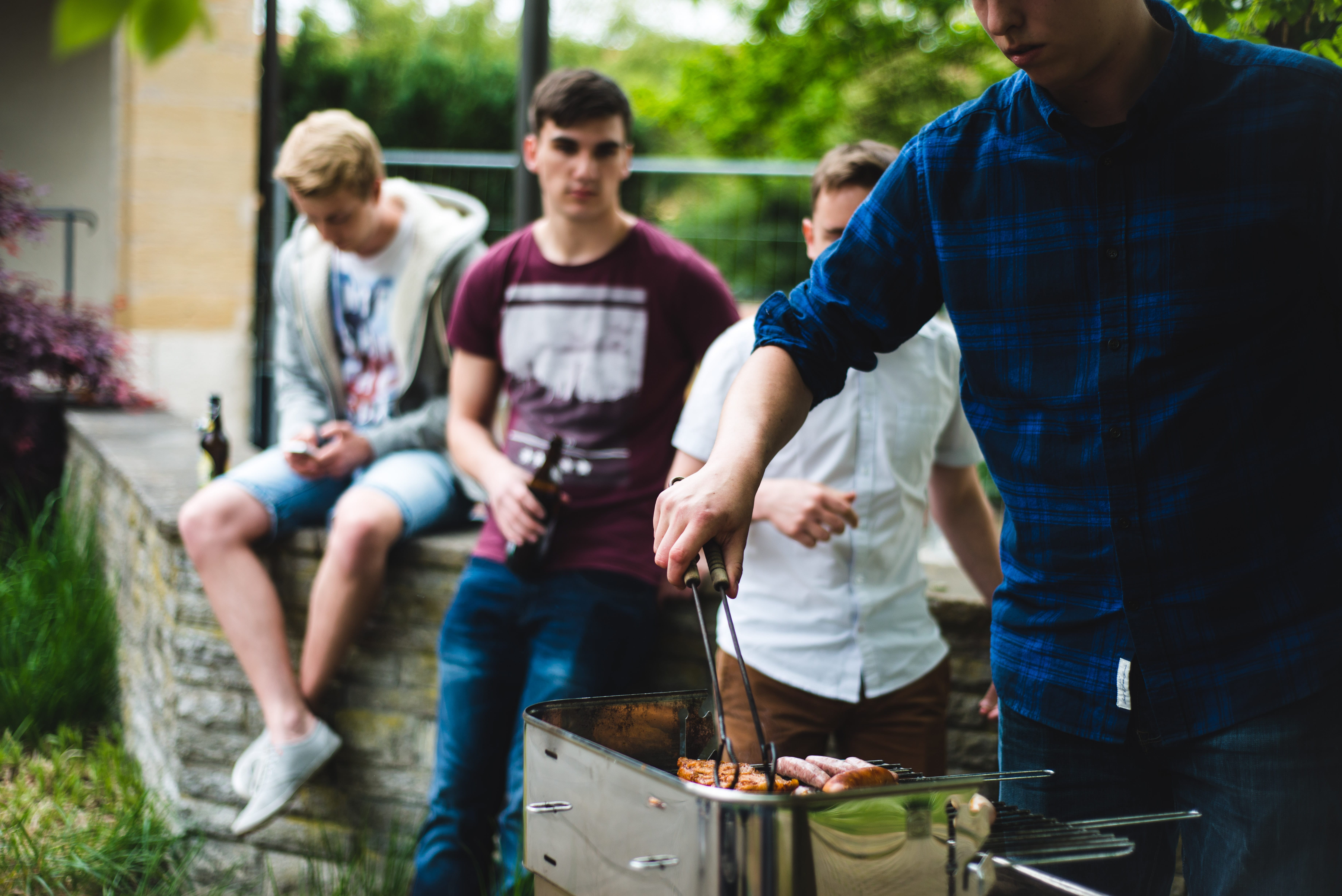 Im Vordergrund steht ein Student am Grill und grillt. Im Hintergrund sitzen drei weitere Studenten mit einem Bier in der Hand auf der Mauer und schauen zum Grill.