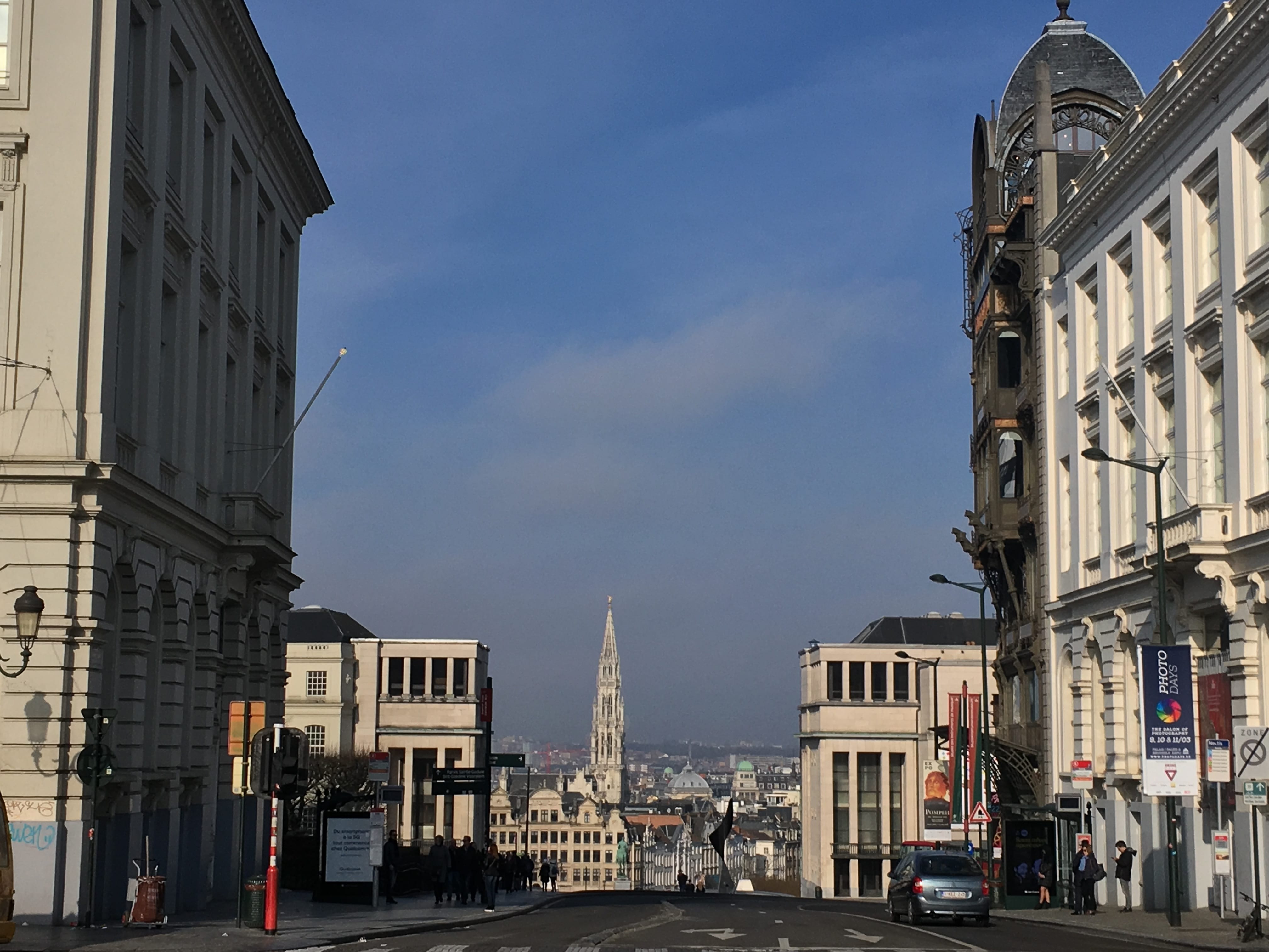 Eine Straße mit Gebäuden in Brüssel. Es ist ein relativ klarer blauer Himmel zu sehen. Links und rechts der Straße befinden sich diverse helle Altbauten. Im Hintergrund sieht man ein Stück der Skyline.