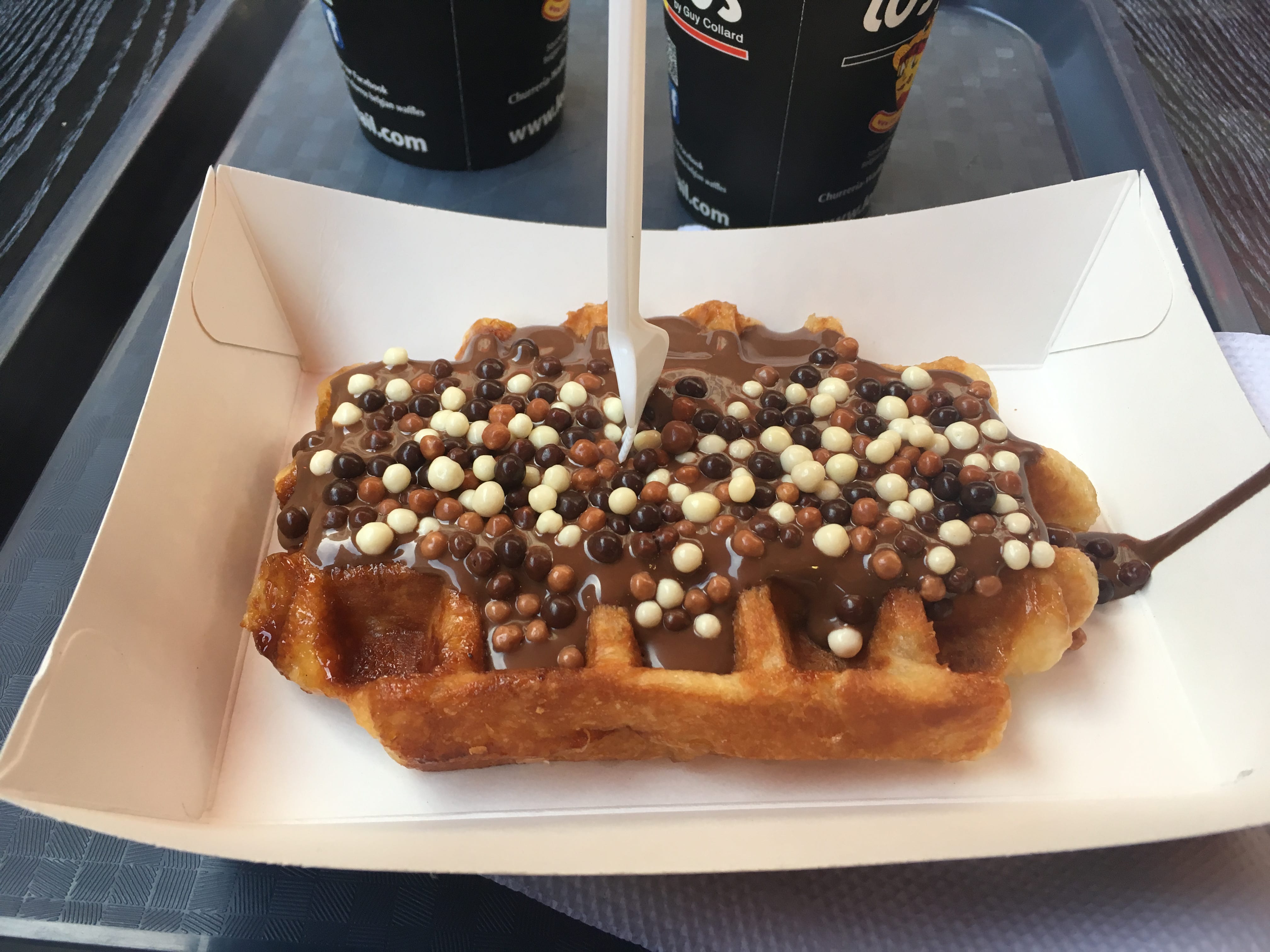 Eine belgische Waffel mit Schokoladensauce und verschiedenfarbigen Schokodrops verziert liegt auf einem Teller.