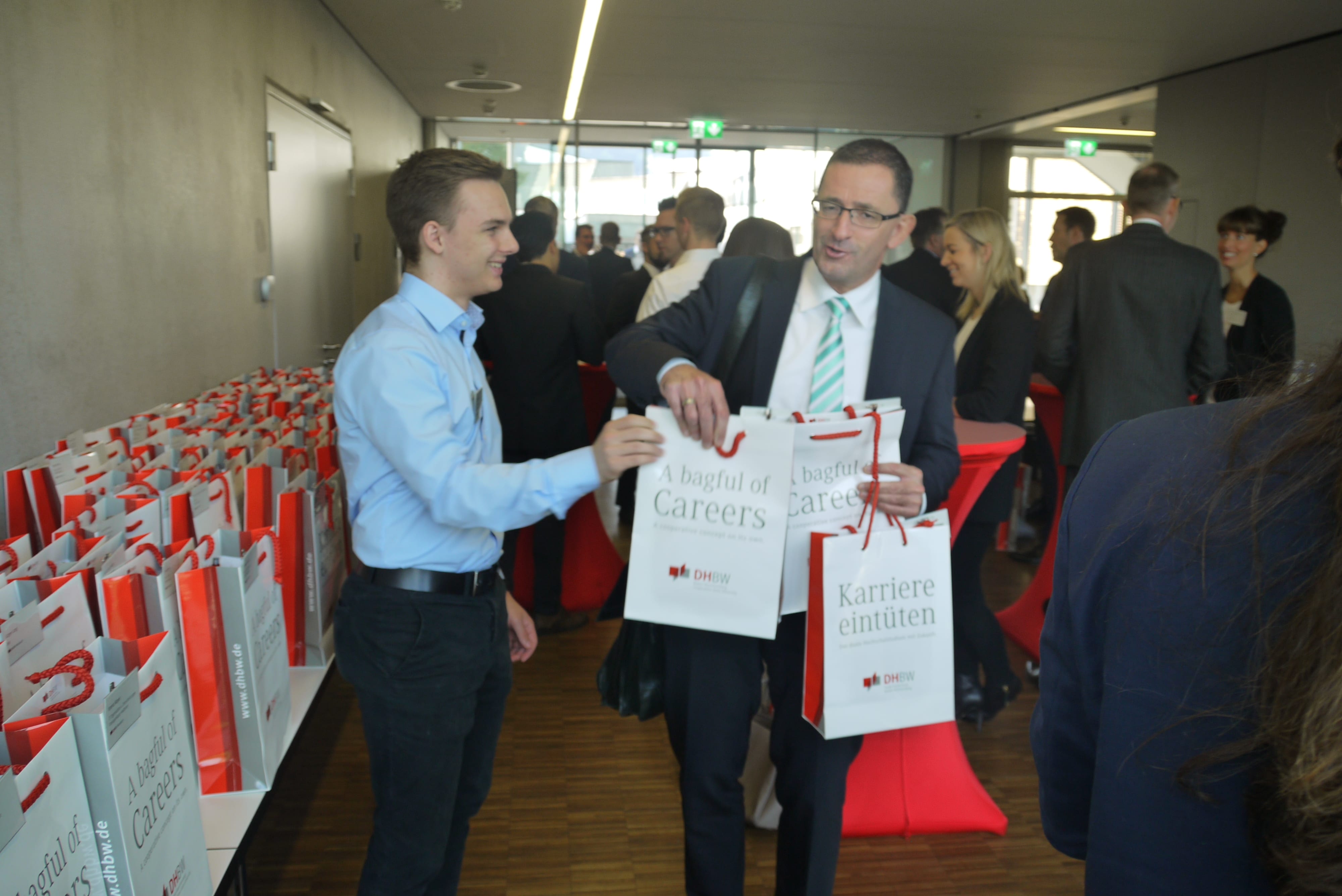 Viele Menschen stehen im Audimax der DHBW Mosbach. Im Vordergrund übergibt ein Mann mehrere Tüten an einen anderen. An der Seite reihen sich auf einem Tisch zahlreiche weitere Tüten.