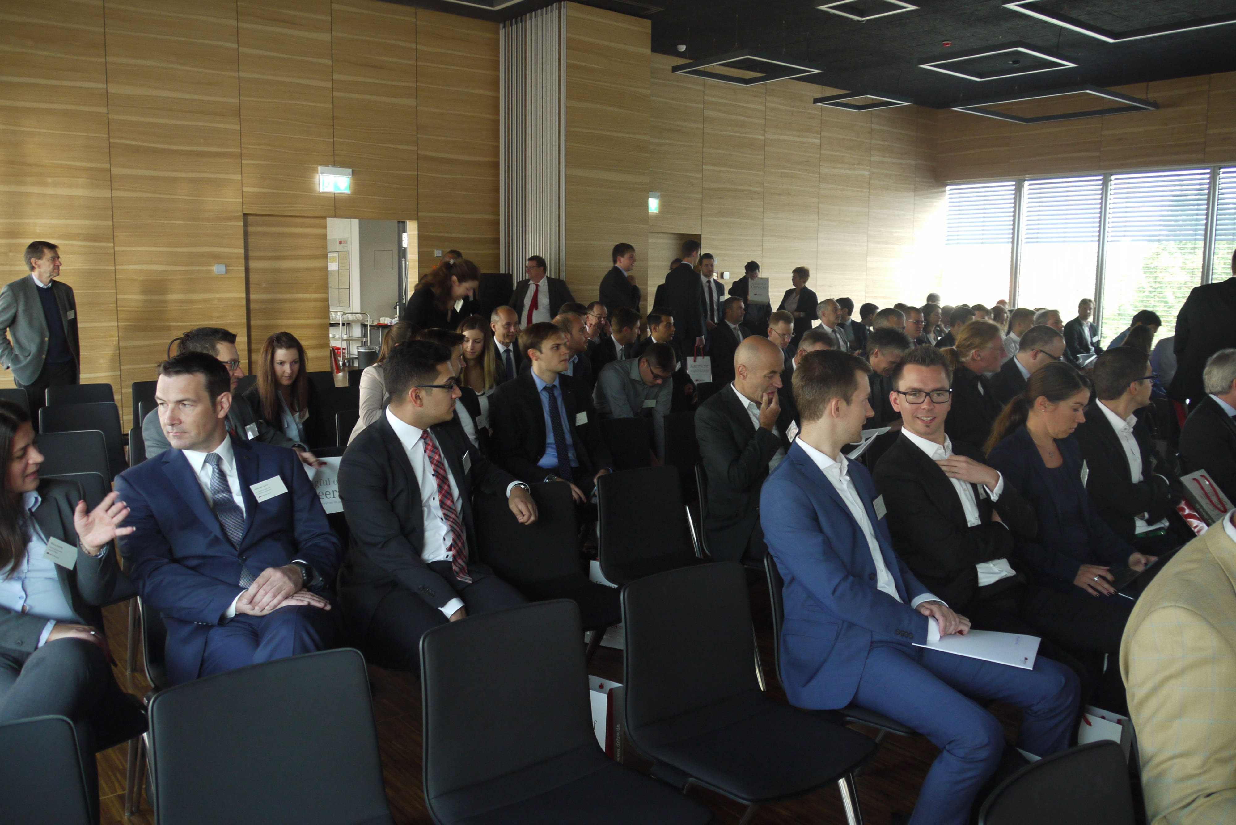 Zahlreiche Menschen sitzten in Anzug auf nebeneinandergereihten Stühlen im Audimax der DHBW Mosbach.