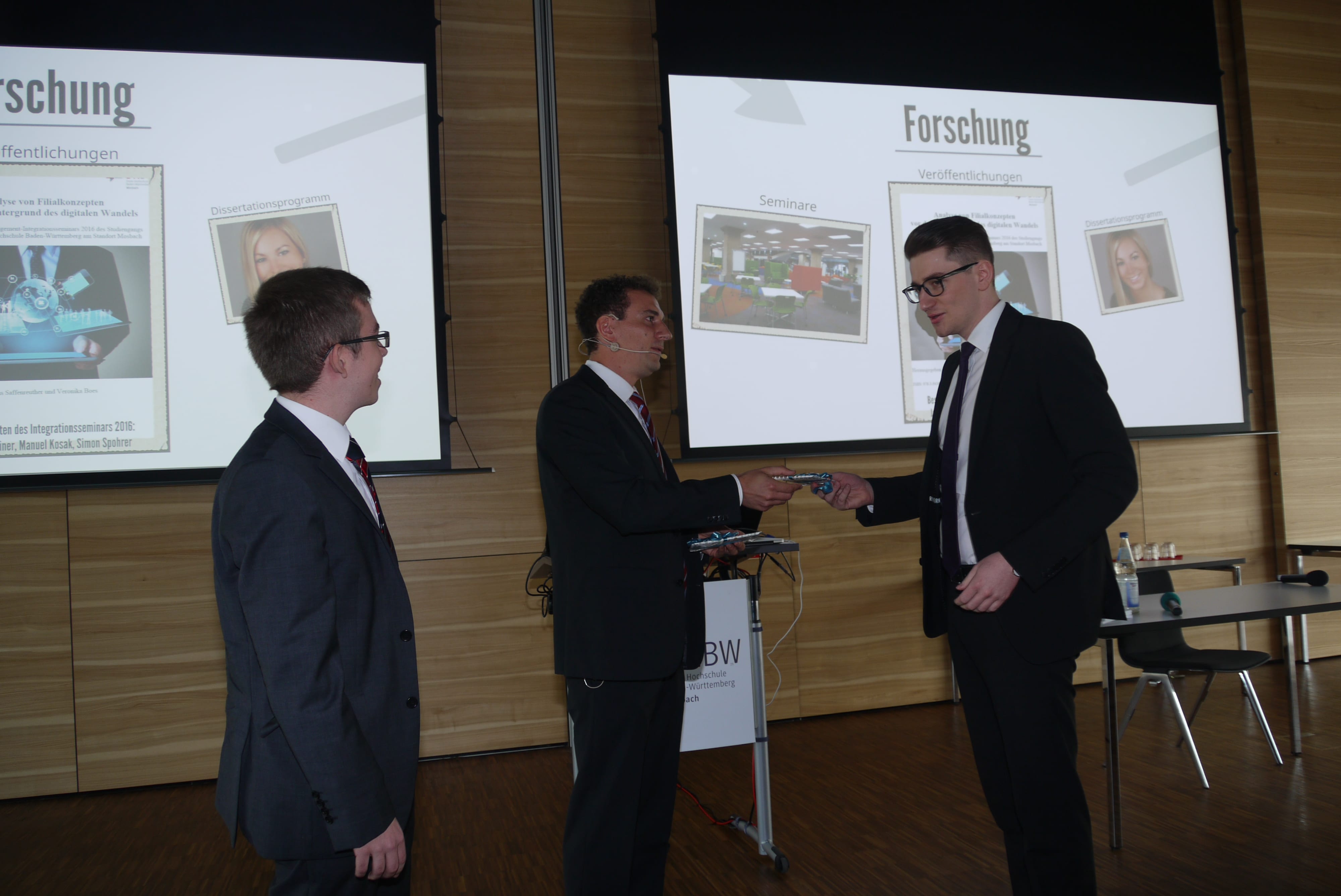 Drei Männer stehen im Anzug im Audimax. Der mittlere überreicht dem rechten Mann etwas. Im Hintergrund sind Folien einer Präsentation zu sehen.