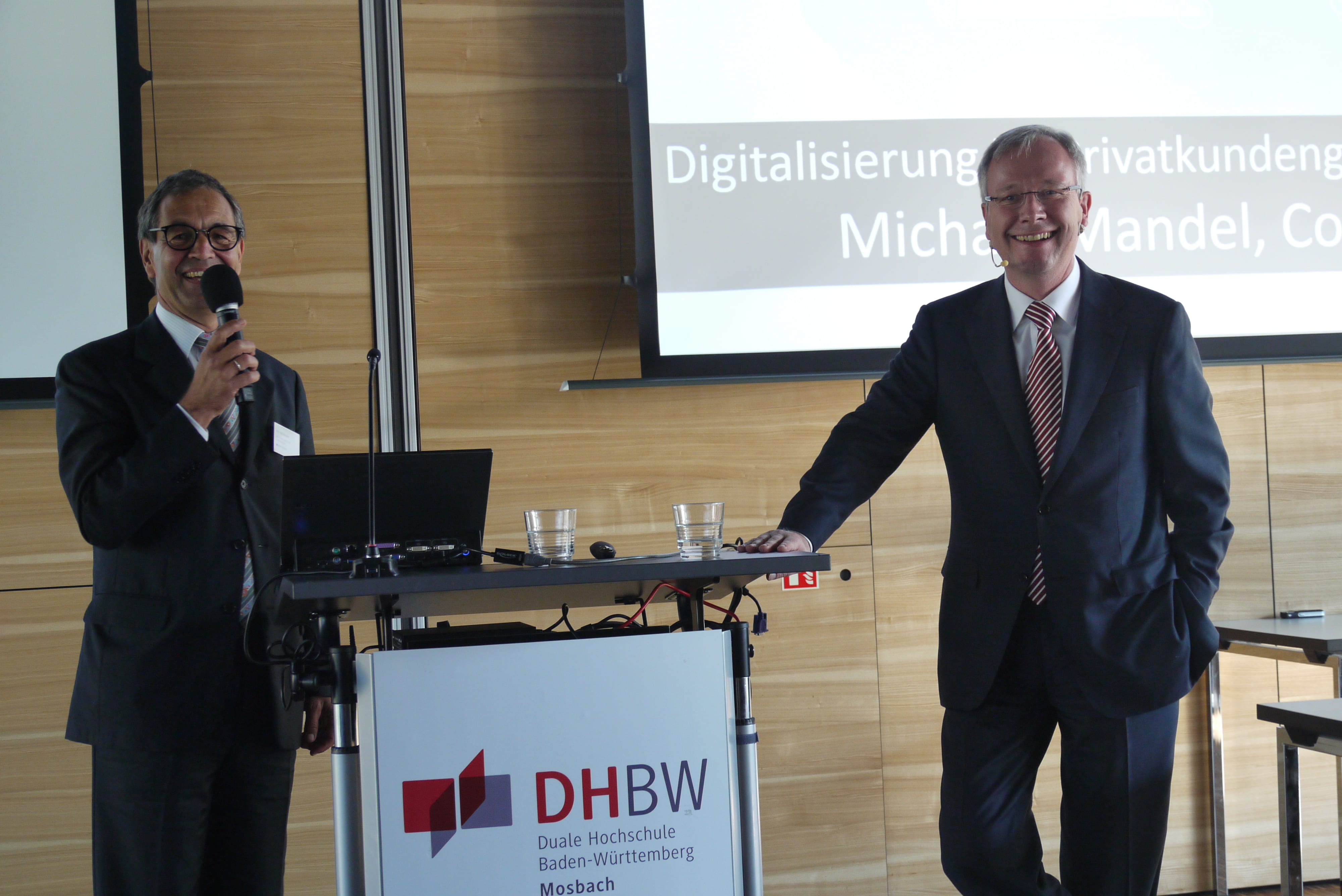 Zwei Männer in Anzügen stehen neben dem Pult im Audimax. Der linke redet in ein Mikrofon.